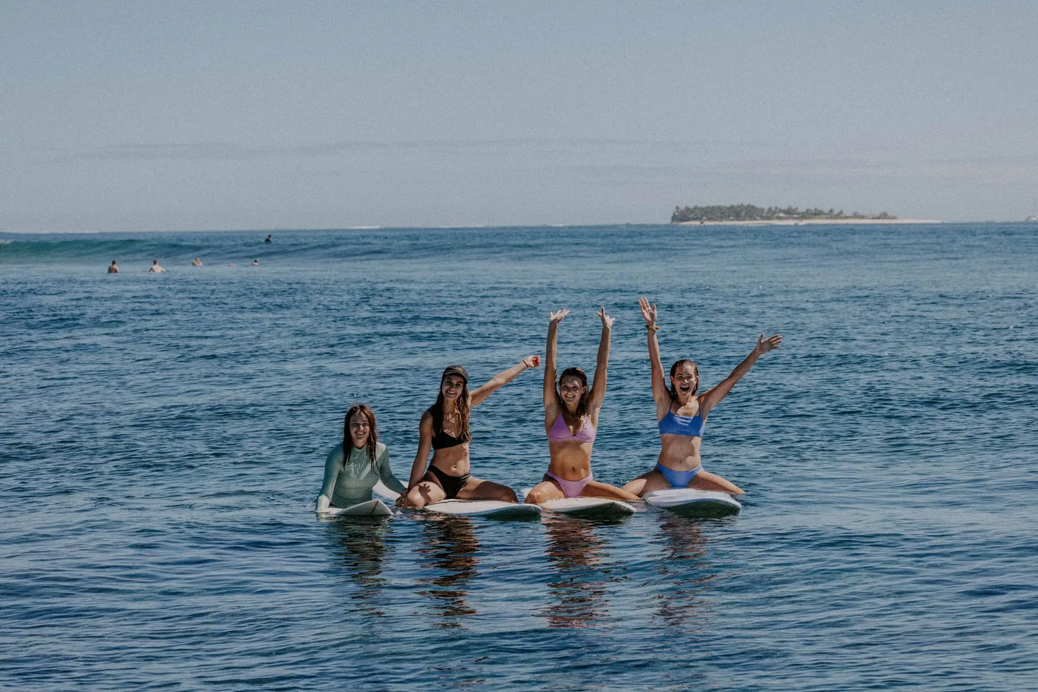 Four women sitting on surfboards in the ocean wearing our bikinis in a range of colours